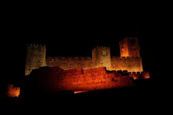 Castelo medieval com torres creneladas iluminado em tons de laranja e amarelo contra fundo escuro noturno.