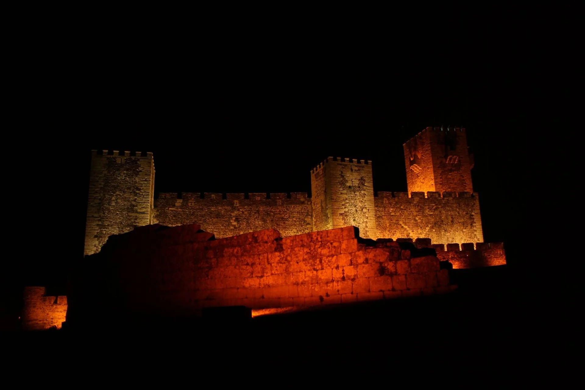 Castelo medieval com torres creneladas iluminado em tons de laranja e amarelo contra fundo escuro noturno.
