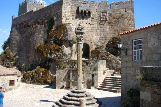Pátio medieval com coluna de pelourinho ao centro, rodeado por muros de pedra e torres do castelo, com vegetação e musgo nas estruturas.