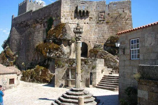 Pátio medieval com coluna de pelourinho ao centro, rodeado por muros de pedra e torres do castelo, com vegetação e musgo nas estruturas.