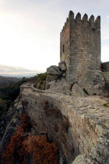 Torre de pedra medieval com ameias, construída sobre rocha granítica, com paisagem montanhosa ao fundo.