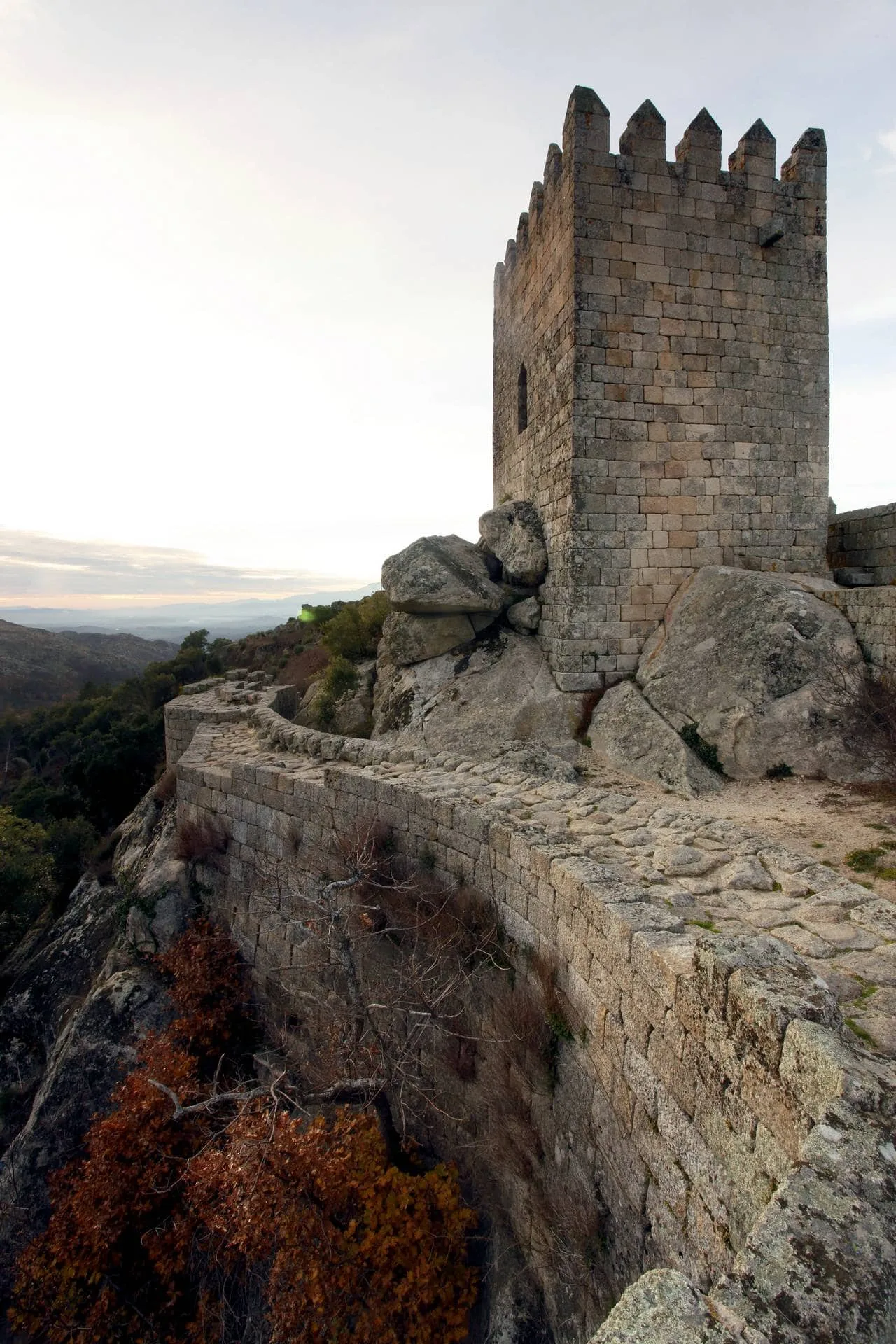 Torre de pedra medieval com ameias, construída sobre rocha granítica, com paisagem montanhosa ao fundo.