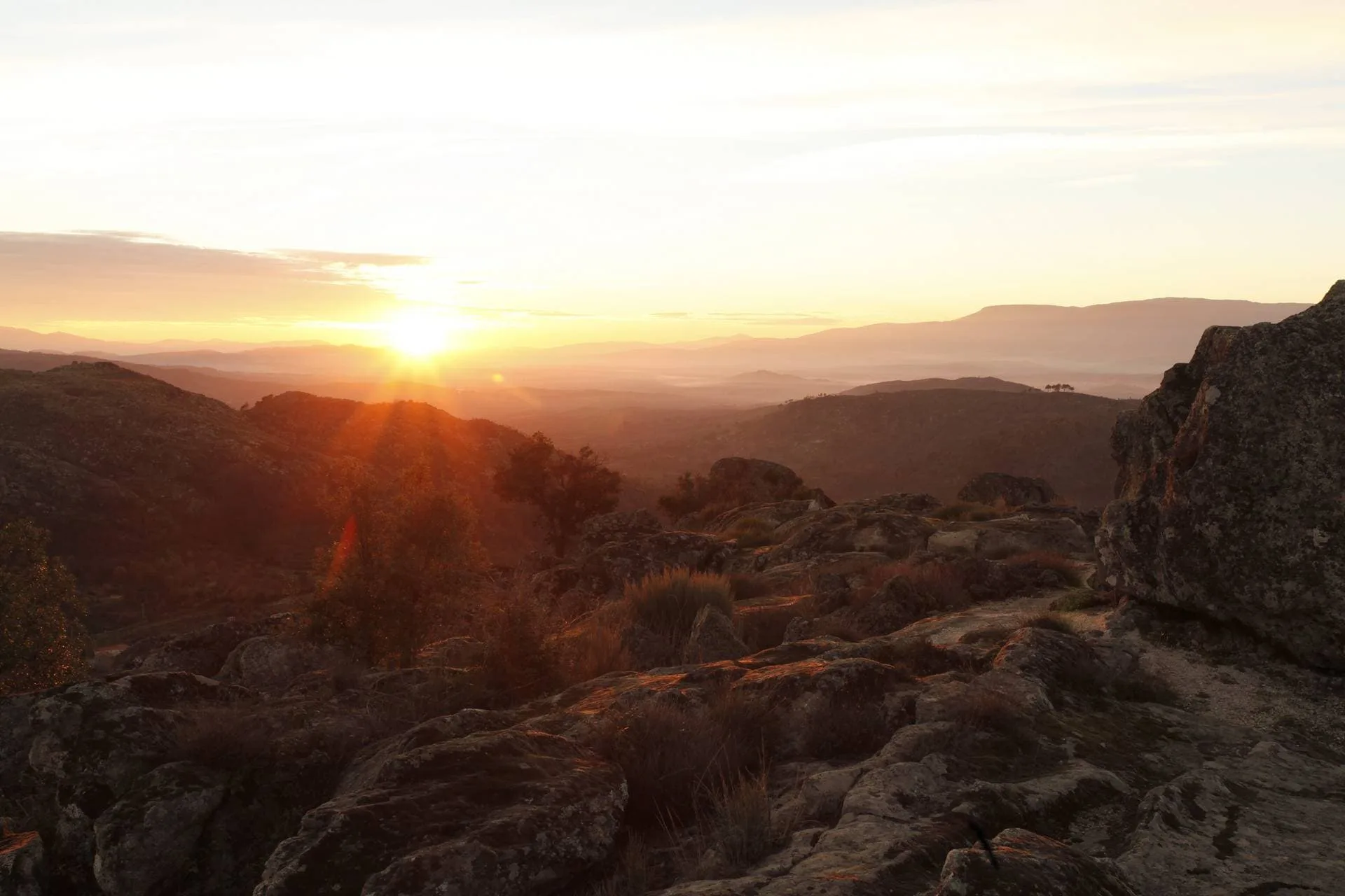 Pôr do sol sobre várias camadas de montanhas e rochedos, com luz dourada iluminando a paisagem árida.