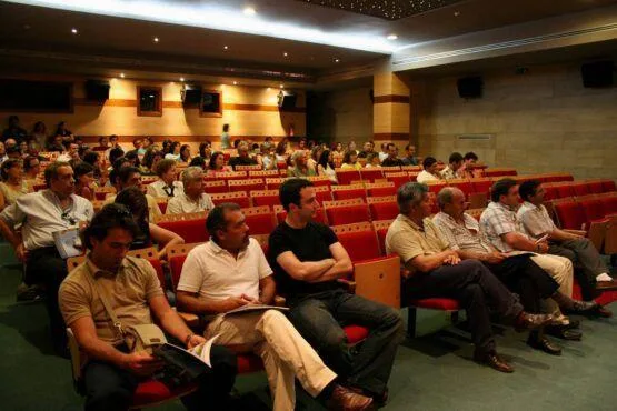 Auditório com centenas de pessoas sentadas em cadeiras vermelhas, assistindo a um evento num espaço com iluminação dourada e paredes de pedra.