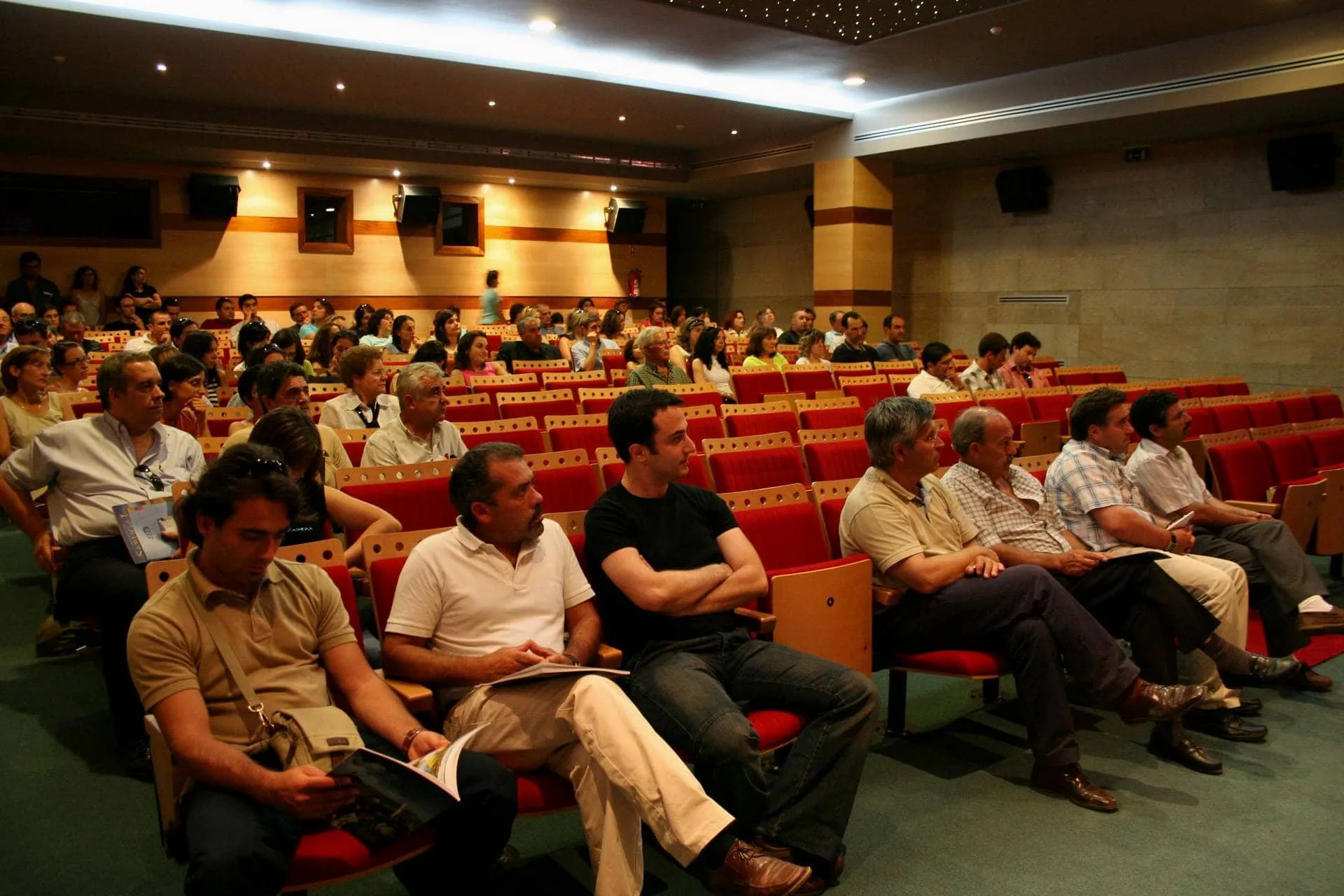Auditório com centenas de pessoas sentadas em cadeiras vermelhas, assistindo a um evento num espaço com iluminação dourada e paredes de pedra.