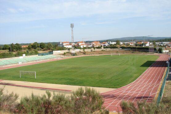 Estádio desportivo com pista de corrida vermelha, campo de futebol verde e piscina descoberta, edifícios residenciais ao fundo.