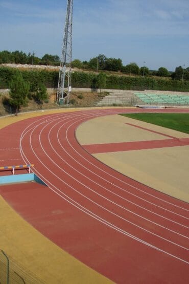 Pista de atletismo com curvas vermelhas e zonas bege, torre de iluminação metálica, bancadas e terreno montanhoso ao fundo.