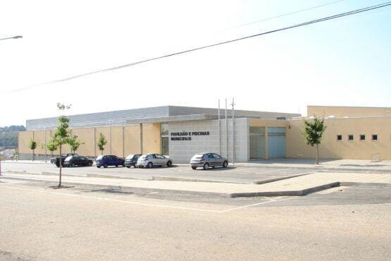 Fachada frontal do Pavilhão e Piscinas Municipais, edifício moderno com painéis amarelos e metalizado, estacionamento com carros, árvores ornamentais.