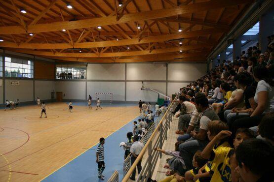 Partida de andebol em curso num pavilhão coberto com estrutura de madeira, crianças no terreno e centenas de espetadores sentados nas bancadas laterais.