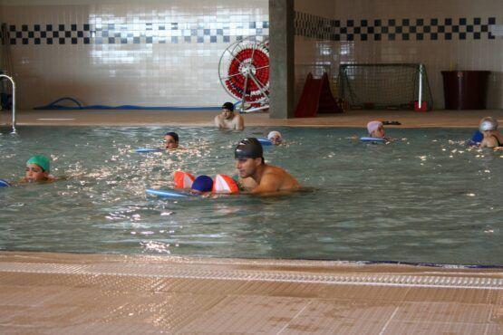Grupo de crianças e instrutor em aula de natação em piscina interior, usando flutuadores e equipamento de segurança.