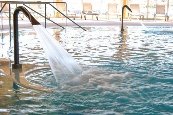 Piscina interior com cascata em funcionamento, espreguiçadeiras de madeira ao fundo e corrimãos metálicos.