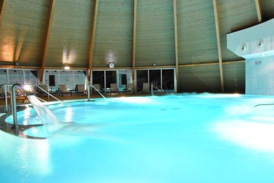 Piscina interior aquecida com água azul-turquesa, cobertura abobadada verde-claro e escadas de acesso metálicas, espreguiçadeiras visíveis ao fundo.