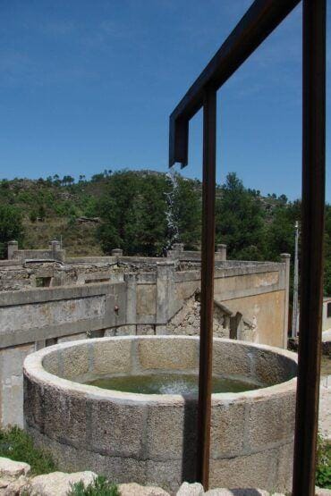 Poço circular de alvenaria com água, enquadrado por estrutura metálica, em ruínas de fortificação com paisagem arborizada ao fundo.