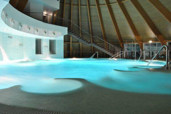 Interior de um complexo termal com piscinas de água quente azul-turquesa, escadas metálicas e estrutura de madeira no tecto.