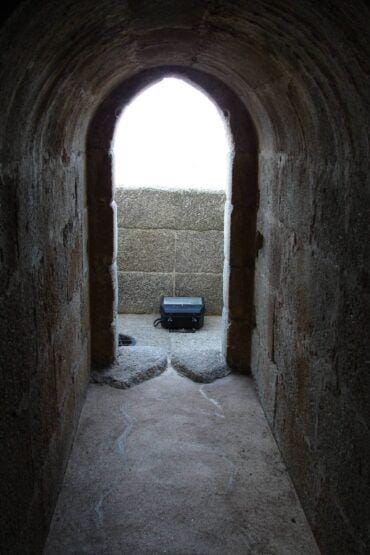 Corredor de pedra em ruínas de fortaleza com arcada apontada, vendo baú azul no espaço interior iluminado