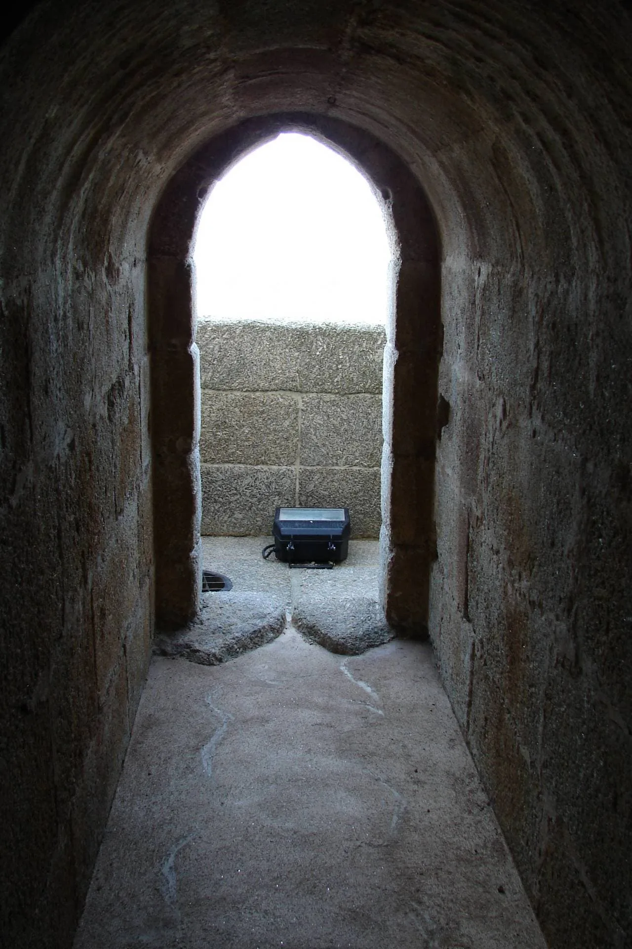 Corredor de pedra em ruínas de fortaleza com arcada apontada, vendo baú azul no espaço interior iluminado