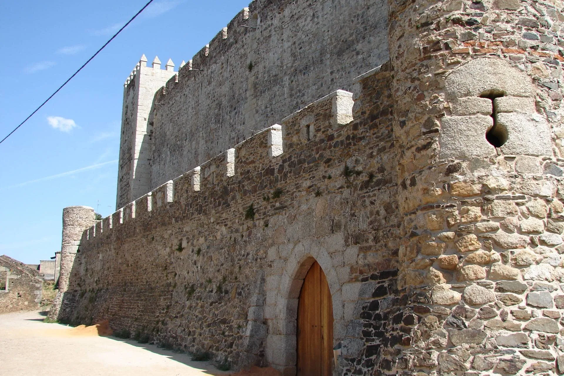 Muralha de pedra com torres almenaradas, porta de arco ogival e abertura circular, sob céu azul claro.