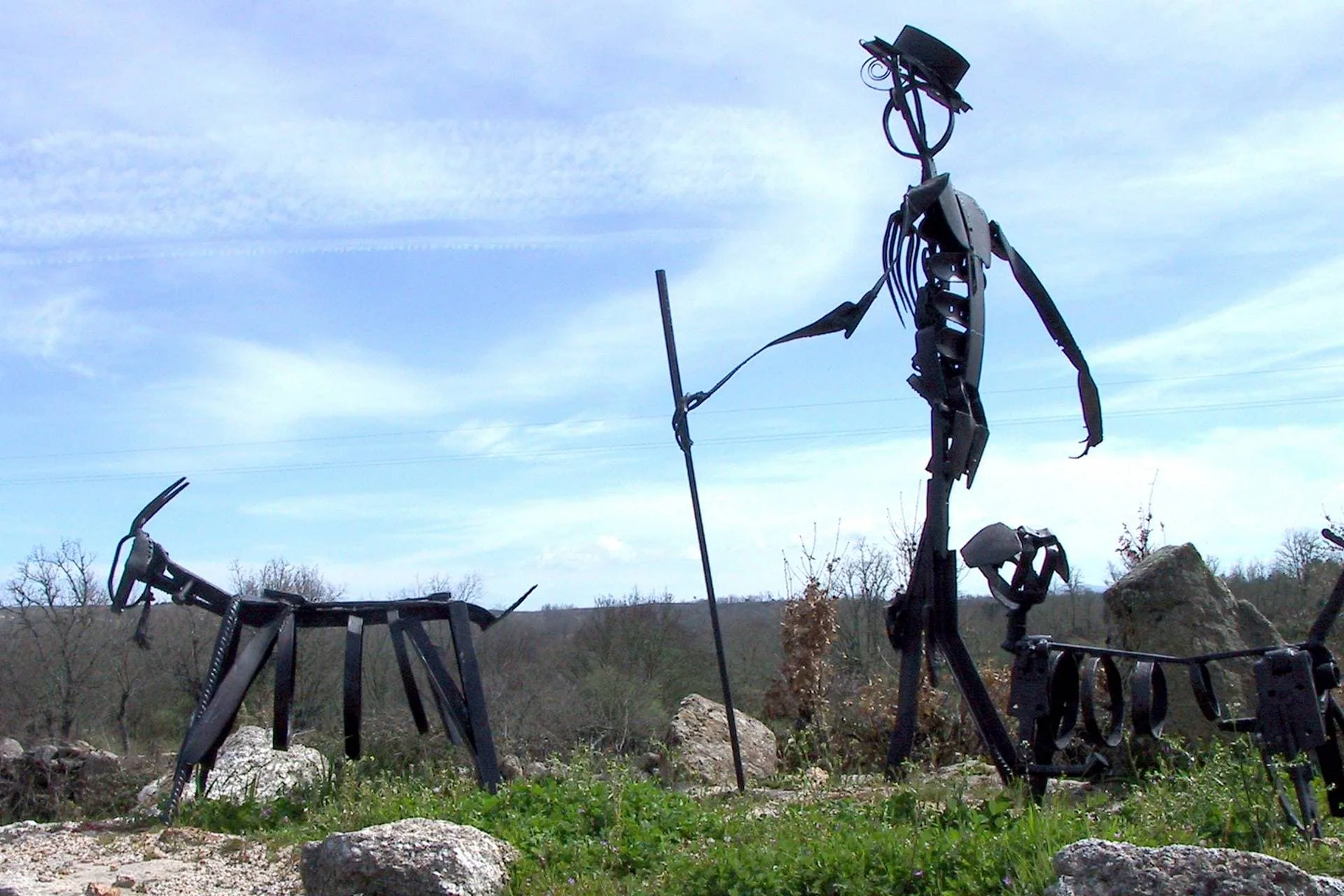 Três figuras humanas de ferro forjado em posições distintas numa paisagem montanhosa com vegetação escassa.