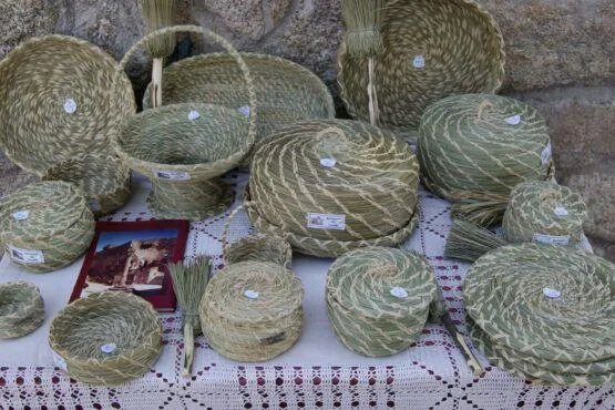 Diversos cestos de palha tecida em tons de verde e natural, com etiquetas brancas, dispostos sobre tecido crochetado junto a pedra.