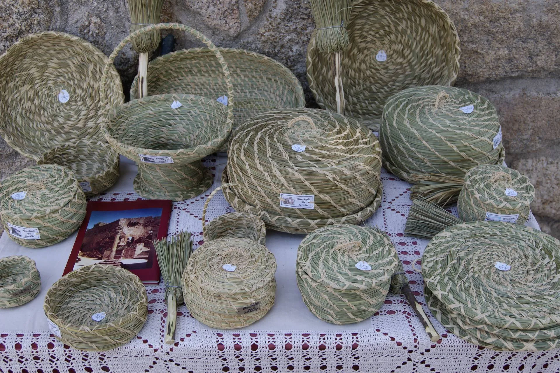 Diversos cestos de palha tecida em tons de verde e natural, com etiquetas brancas, dispostos sobre tecido crochetado junto a pedra.
