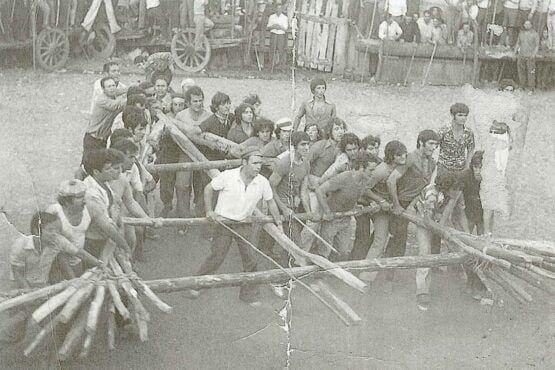 Grupo de pessoas, adultos e crianças, participa em jogo de corda num espaço aberto com espectadores ao fundo.