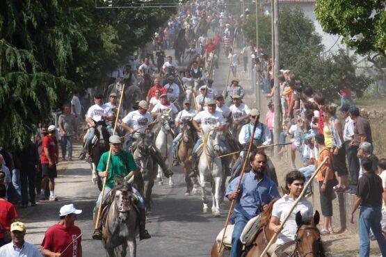 Grupo de cavaleiros a cavalo numa estrada rural, rodeados por centenas de pessoas a pé ao longo de ambos os lados da via.
