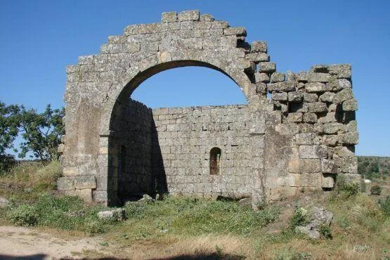Estrutura medieval em ruína com grande arco de pedra, paredes de pedra irregular e pequena abertura rectangular, cercada por vegetação e colinas ao fundo.