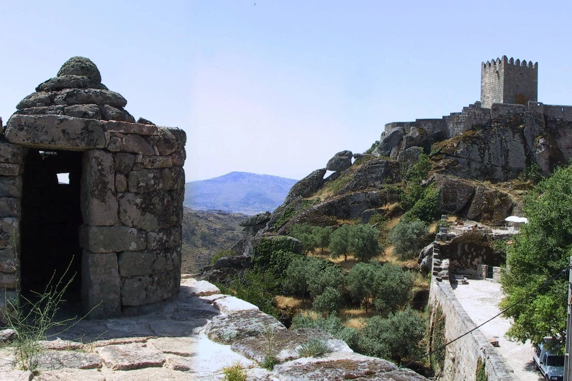 Ruínas de fortificação medieval com torre almoenada ao fundo, estrutura cilíndrica em primeiro plano e paisagem montanhosa de fundo.