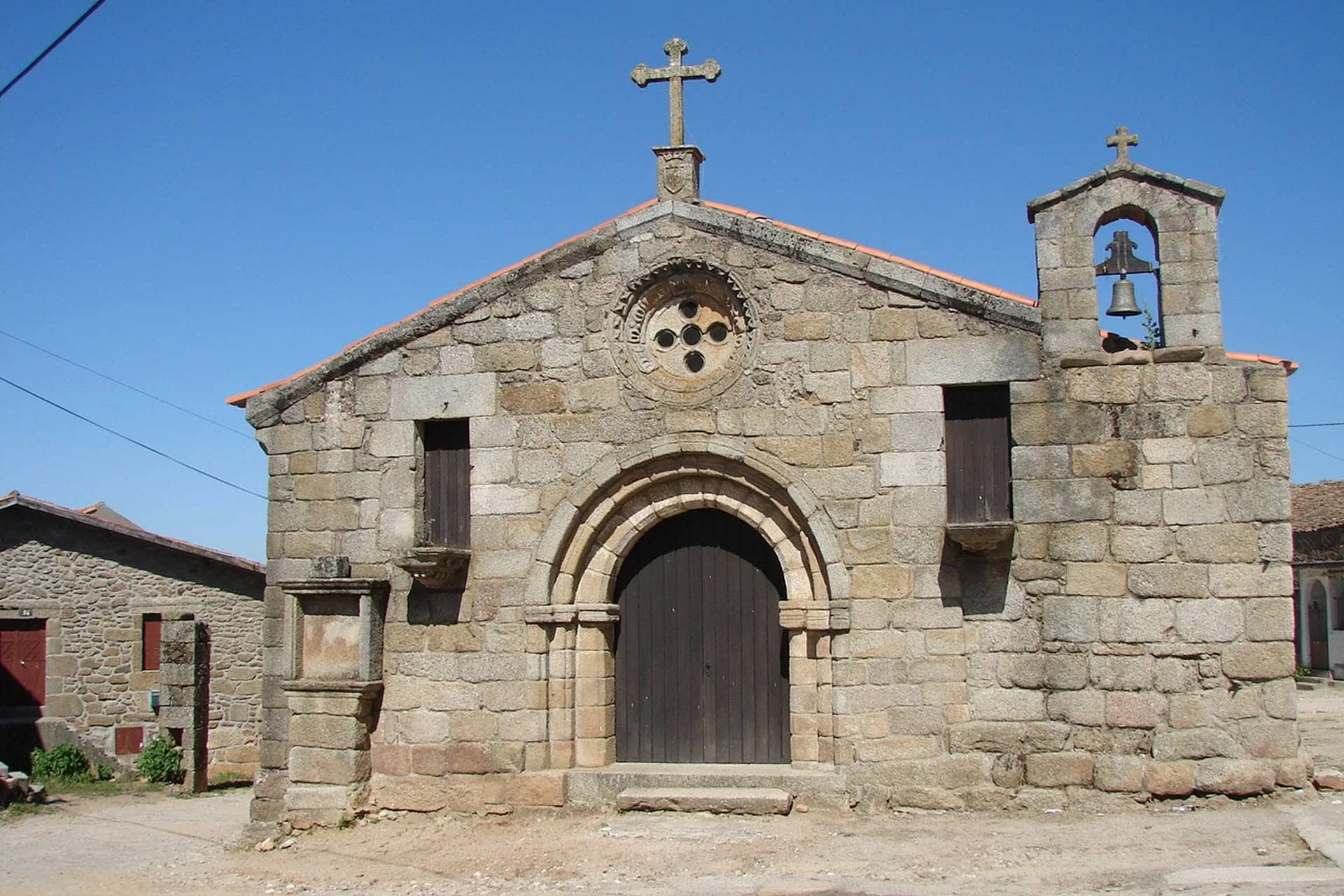 Capela de pedra com fachada Românica, cruz no telhado, janelas pequenas, porta arqueada escura e sino na torre lateral direita.