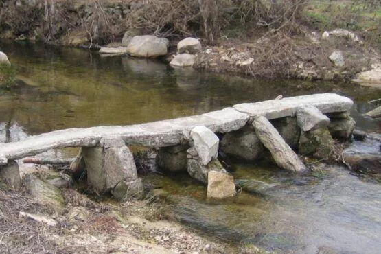 Passagem construída com tronco e pedras sobre ribeira com vegetação degradada ao fundo.
