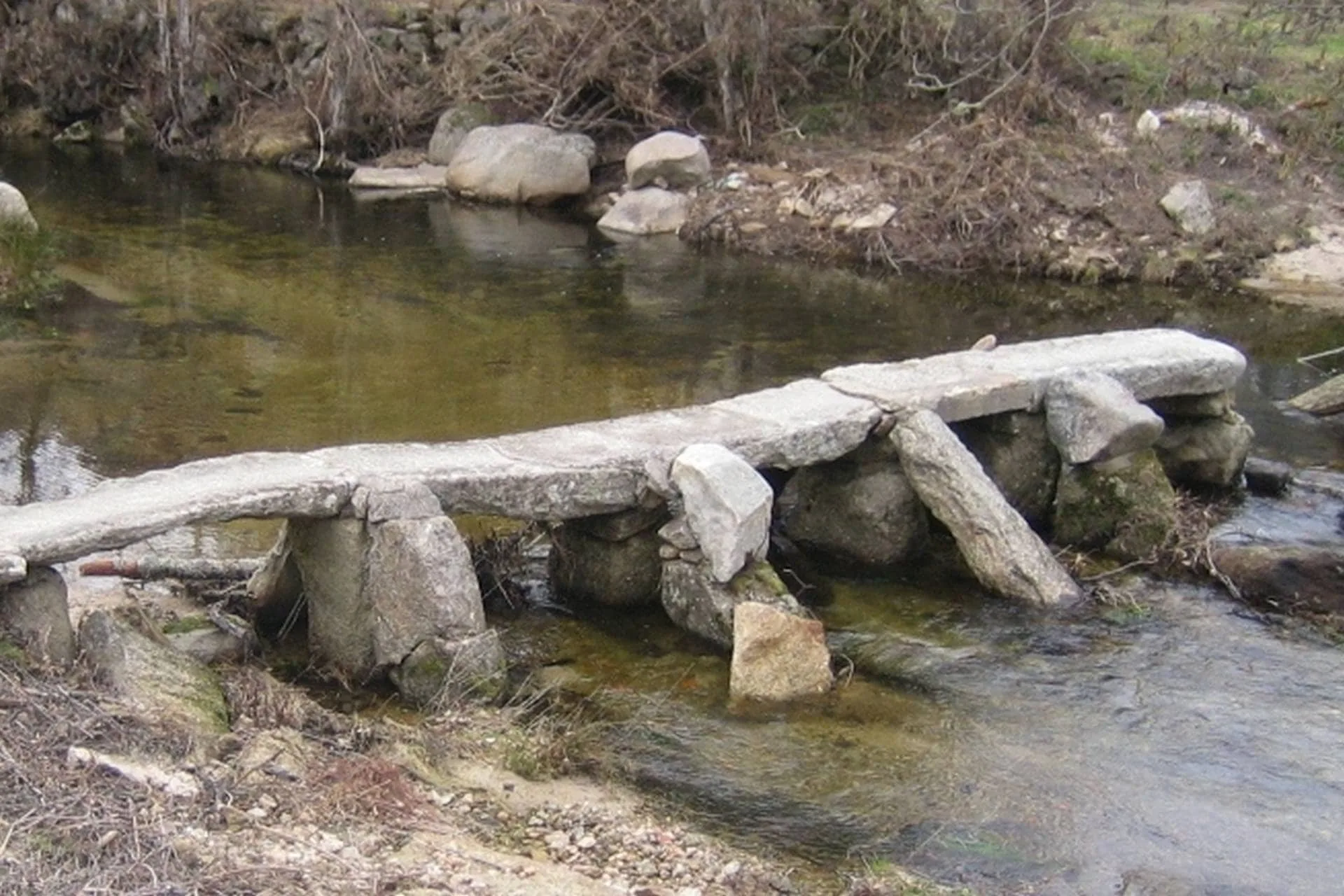 Passagem construída com tronco e pedras sobre ribeira com vegetação degradada ao fundo.