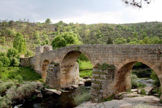 Ponte de pedra com três arcos sobre rio, rodeada de vegetação e colinas arborizadas.