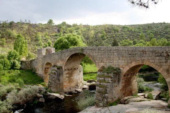 Ponte de pedra com três arcos sobre rio, rodeada de vegetação e colinas arborizadas.