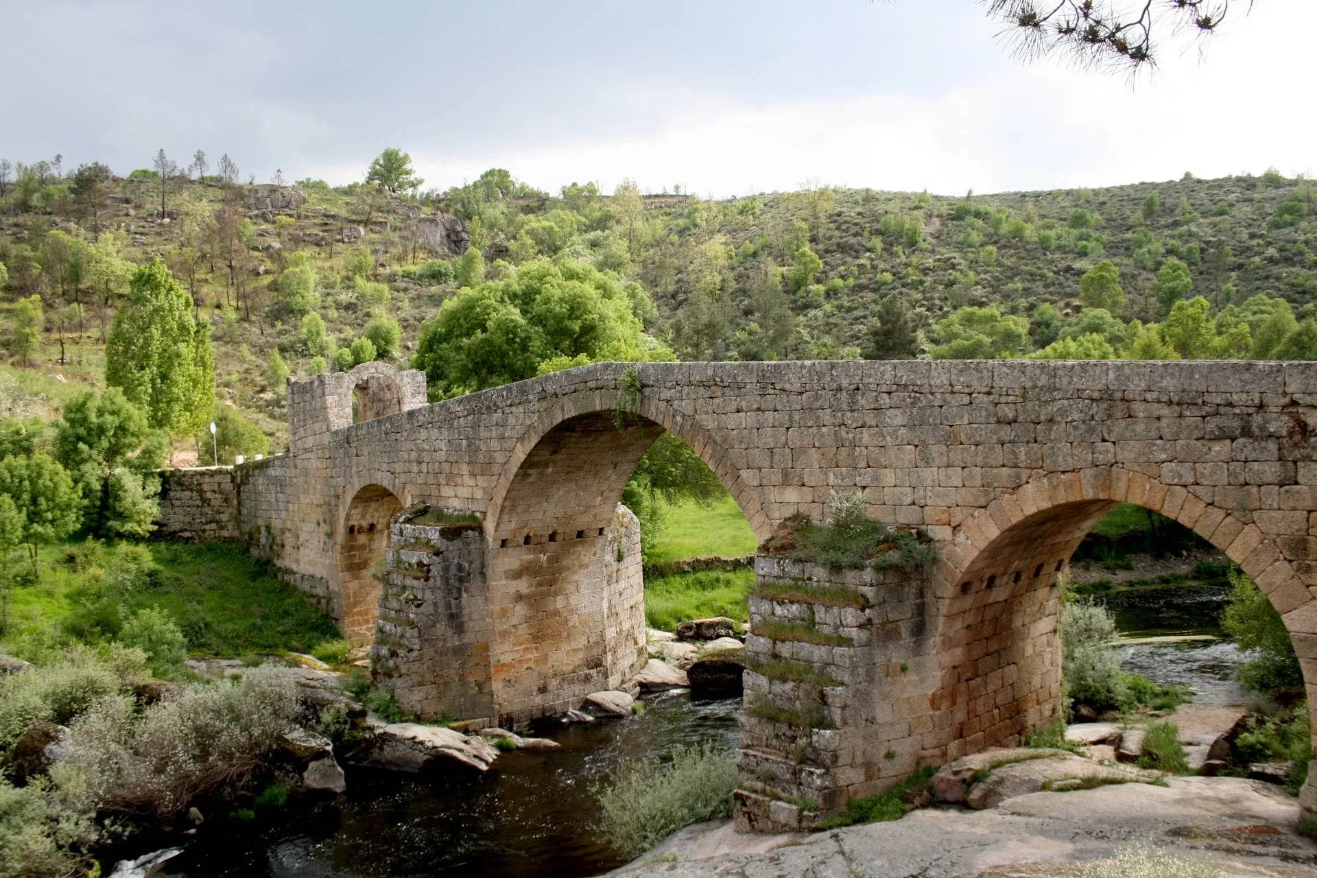 Ponte de pedra com três arcos sobre rio, rodeada de vegetação e colinas arborizadas.