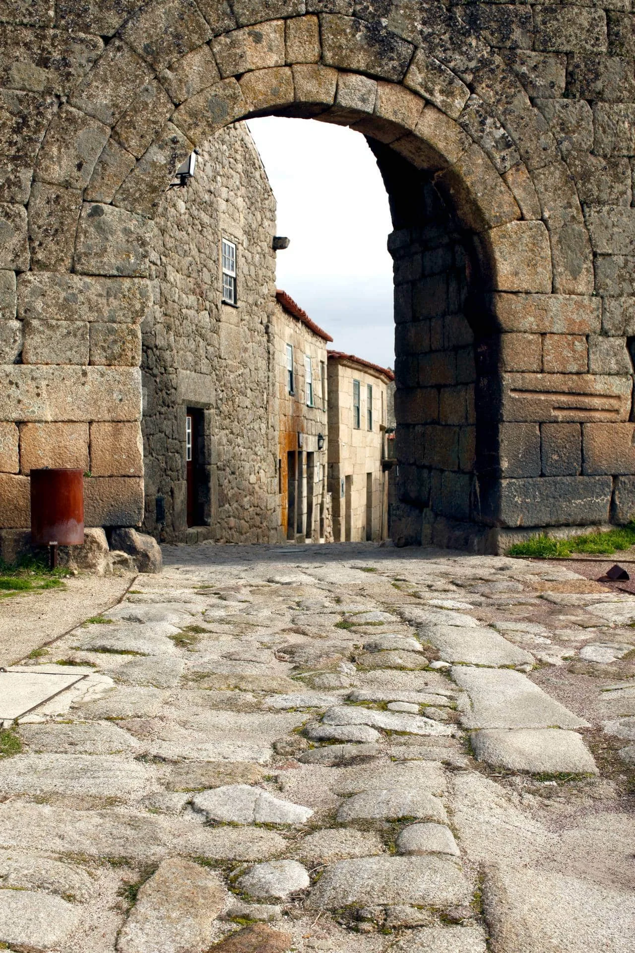 Arco de pedra medieval com abertura central, ladeado por construções antigas de alvenaria em pedra.