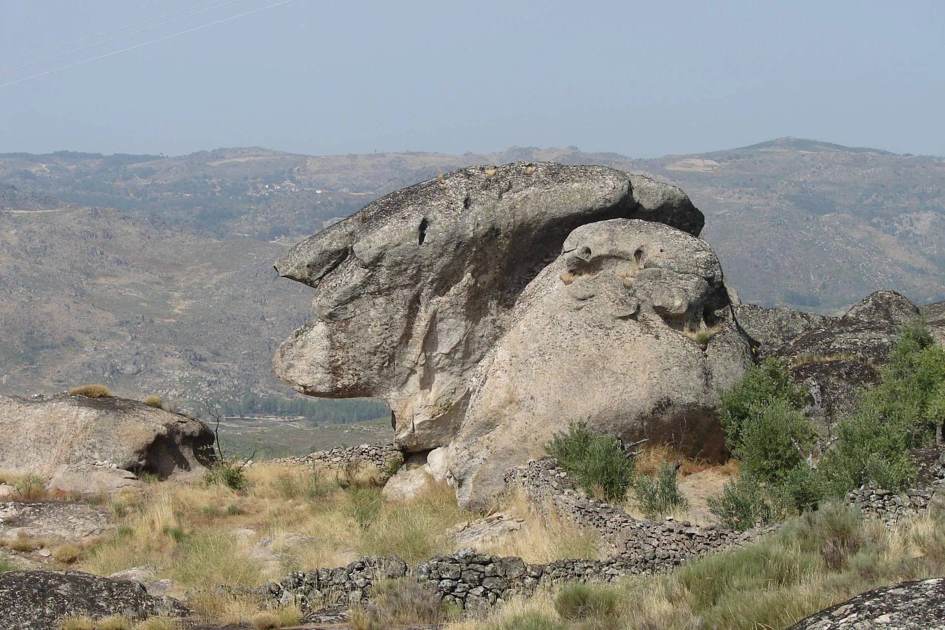 Formação rochosa granítica com aparência zoomórfica numa paisagem montanhosa beirã, com várias elevações ao fundo.