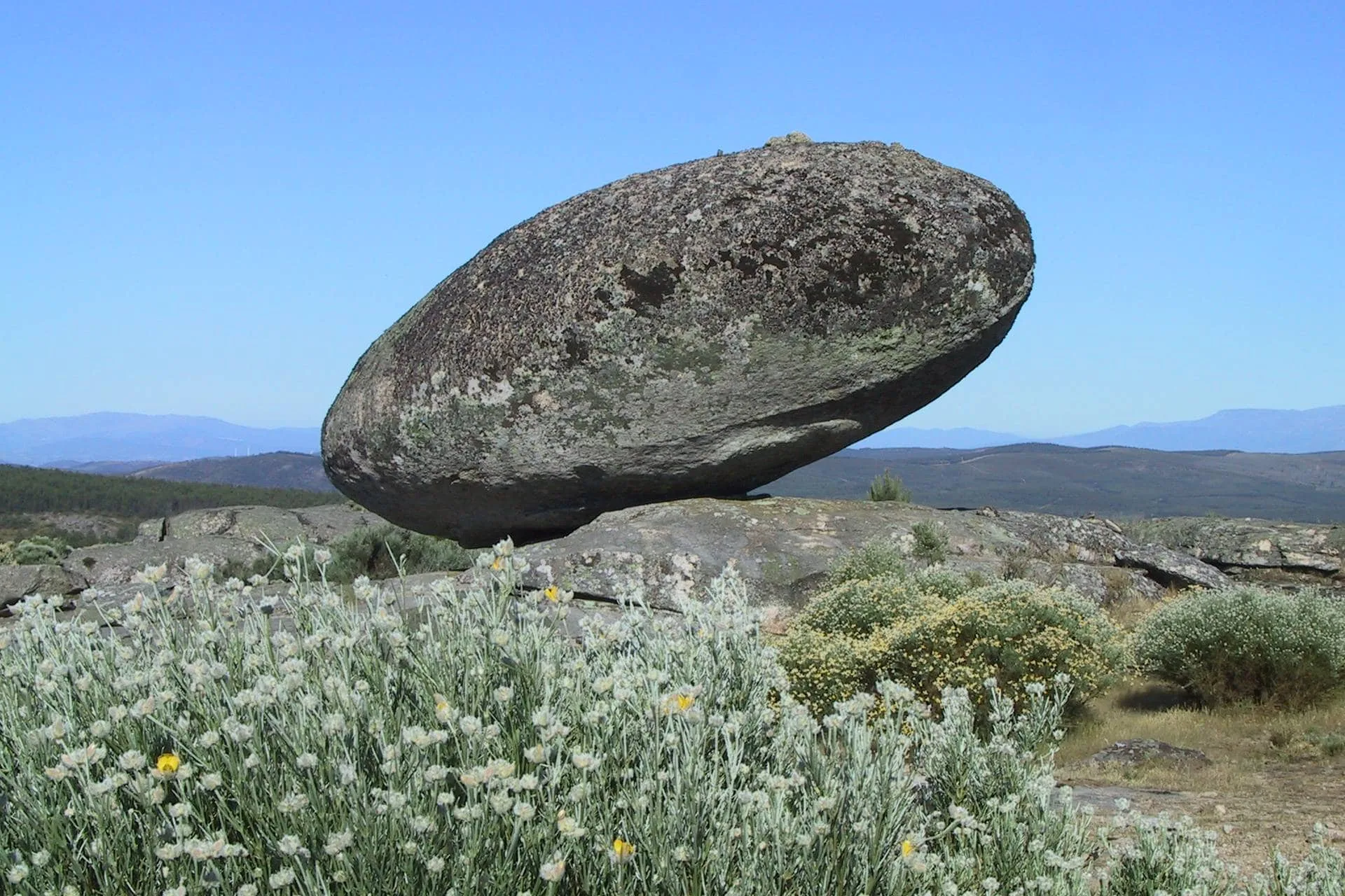 Rocha arredondada equilibrada sobre afloramento rochoso em paisagem árida com vegetação dispersa e montanhas ao fundo.
