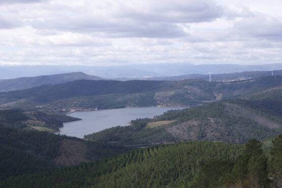 Vale montanhoso com floresta de coníferas, albufeira central e aerogerador visível no horizonte.