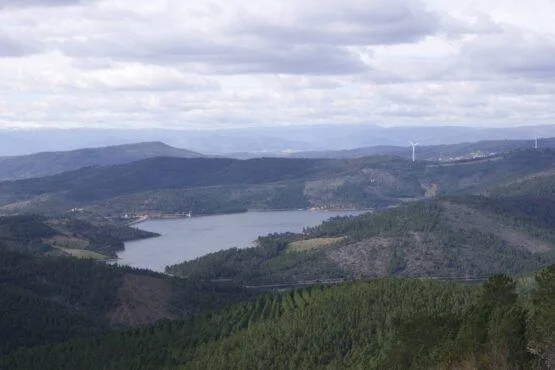Vale montanhoso com floresta de coníferas, albufeira central e aerogerador visível no horizonte.