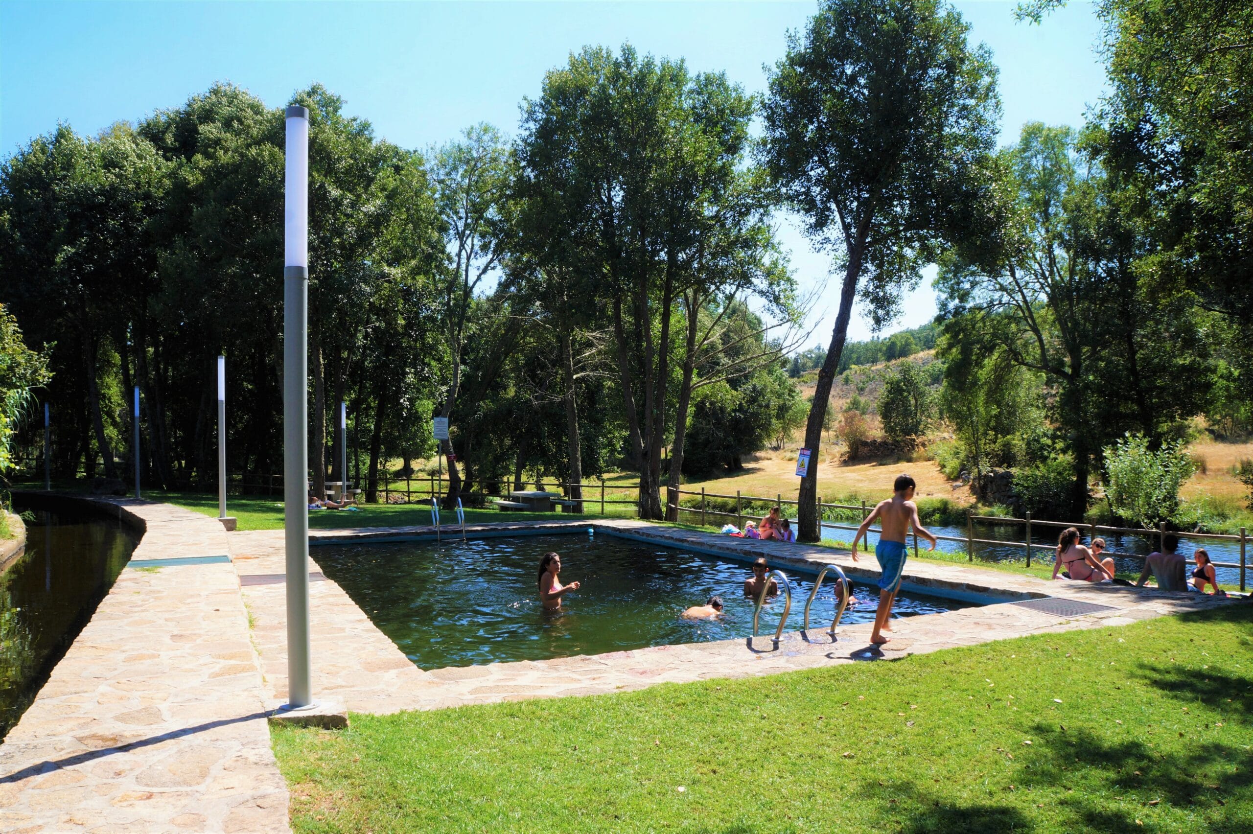 Piscina natural junto a rio, rodeada de árvores, com várias crianças e adultos a nadar e brincar na água.
