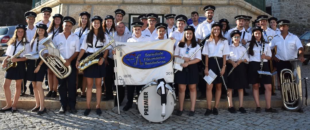 Banda de música com aproximadamente 40 músicos em uniforme branco e preto, segurando instrumentos de sopro, posando com estandarte da Sociedade Filarmónica Pendozense.