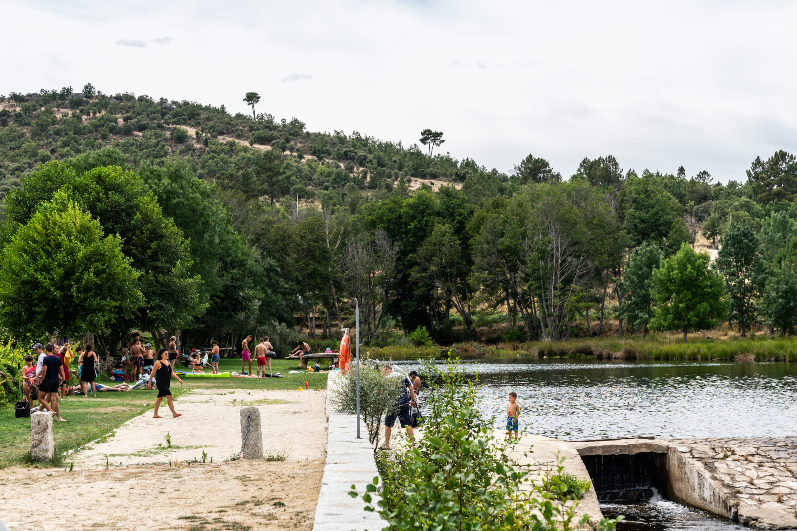 Zona fluvial com praia de areia, pessoas em repouso e recreio, rio com vegetação ribeirinha e colina arborizada ao fundo.