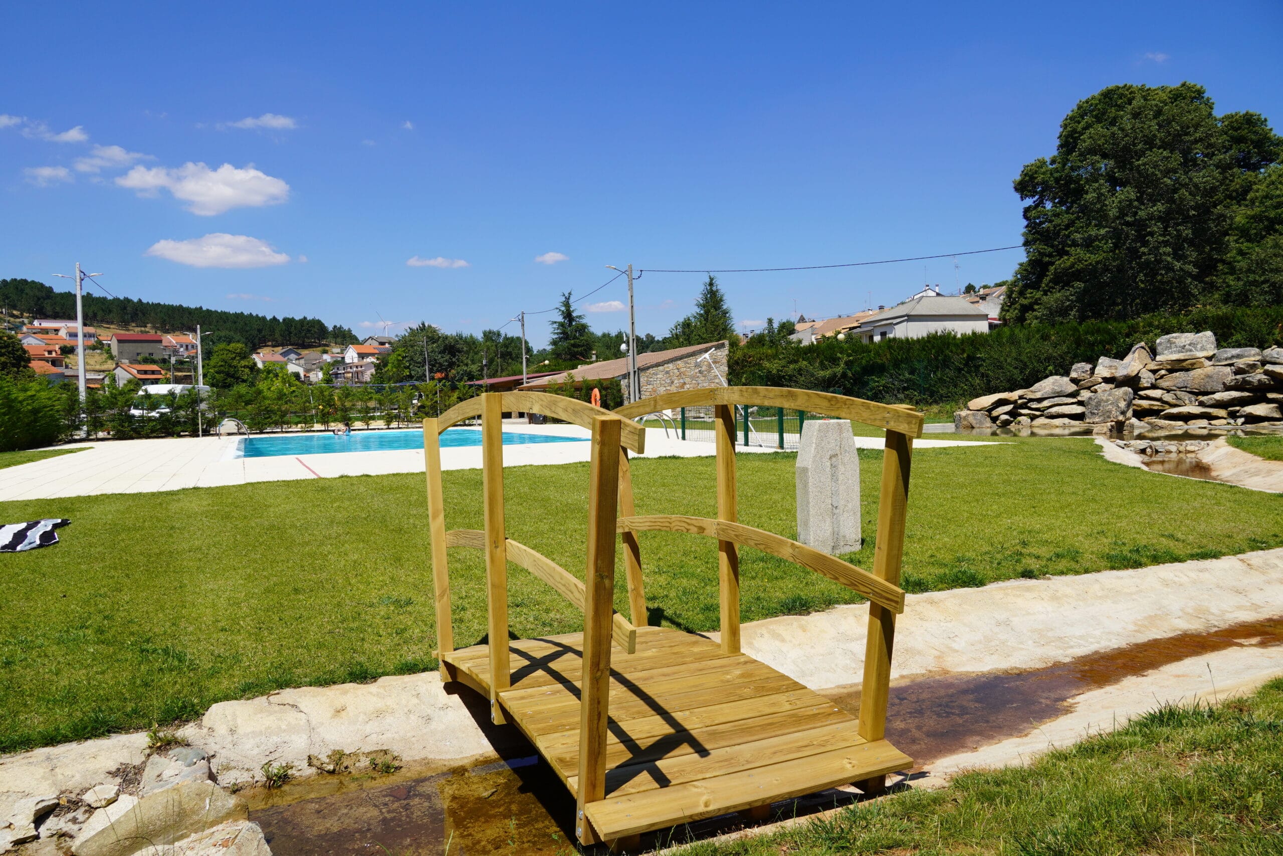 Ponte de madeira rústica sobre pequeno curso de água, com piscina e zona de relva ao fundo numa zona de lazer rural.