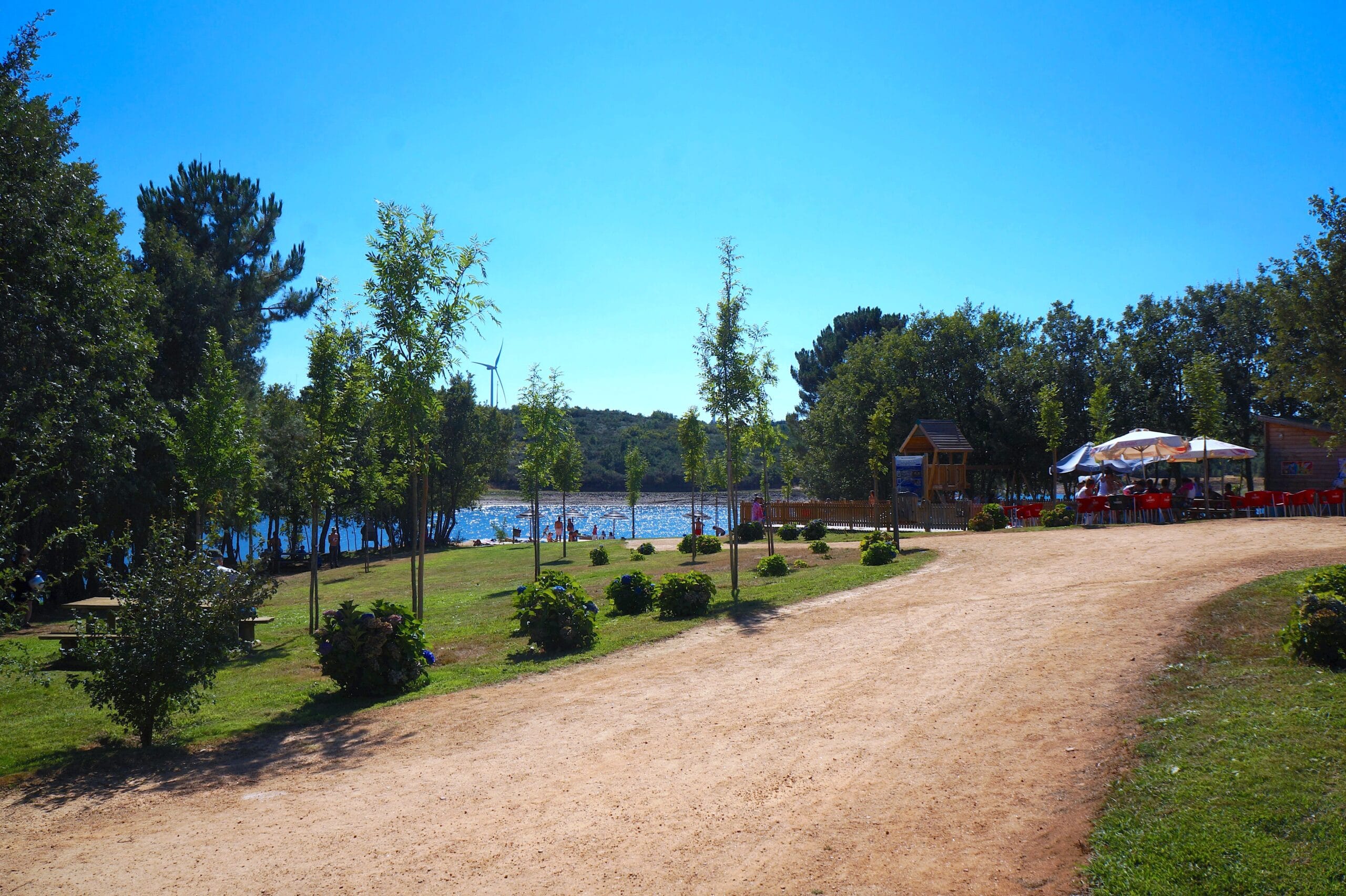 Parque de lazer à beira de rio com piscina azul, zona de areia, espaços verdes e estruturas de acolhimento sob céu limpo.