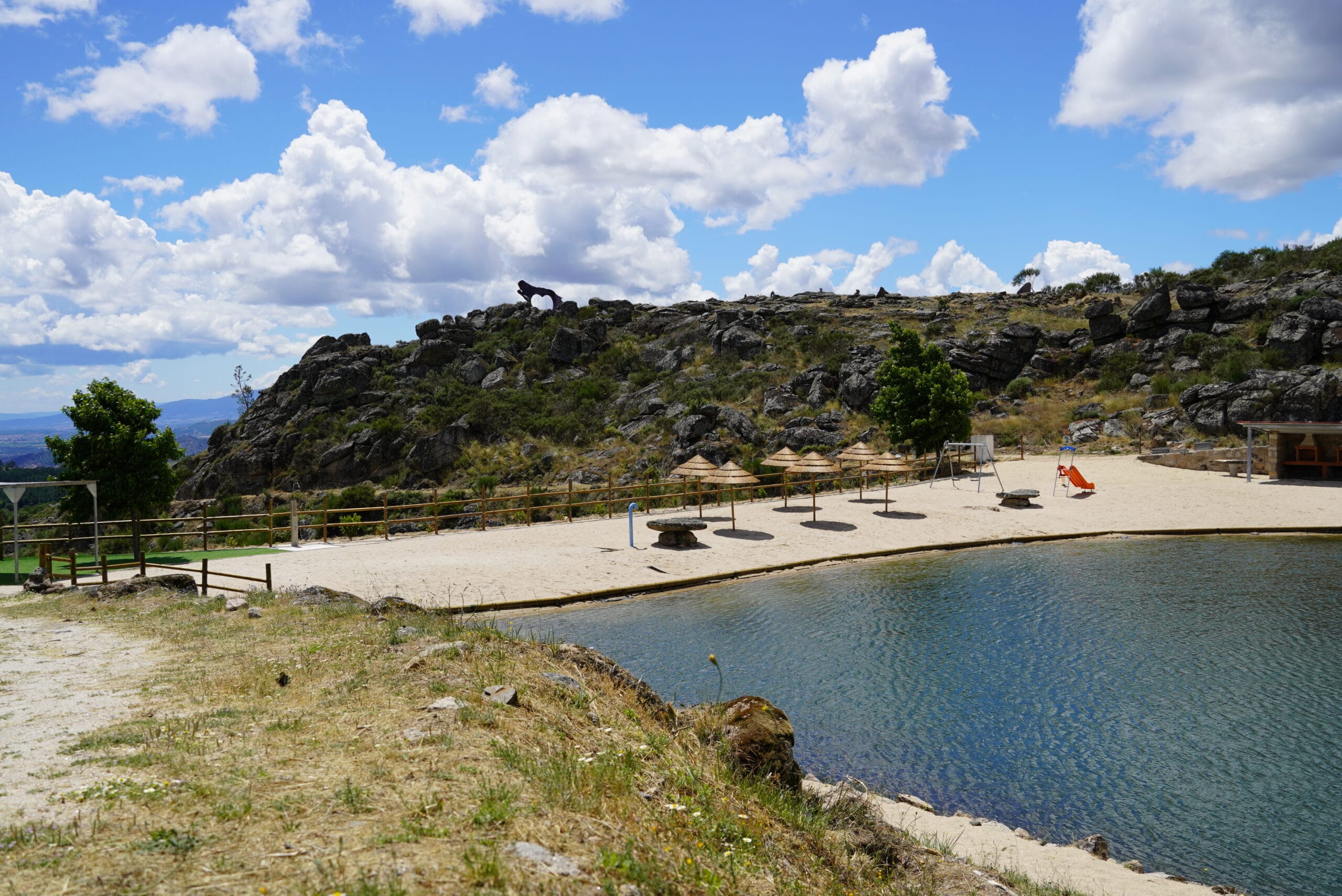 Zona de lazer junto a água com parque infantil, estruturas de sombra e colina rochosa ao fundo, sob céu parcialmente nublado.