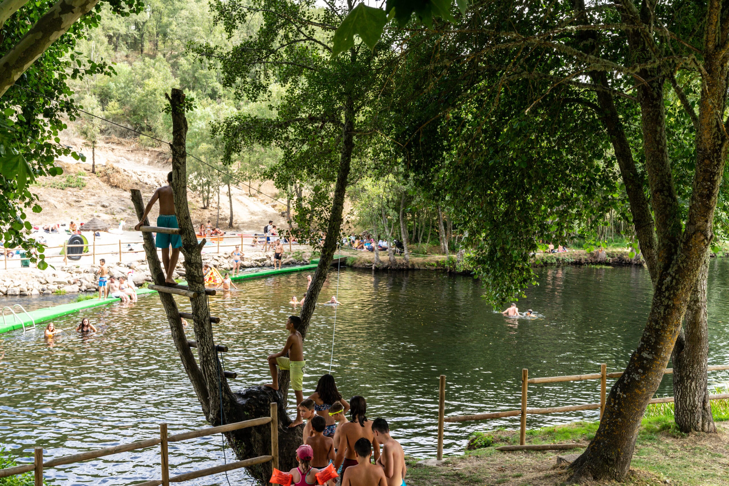 Zona de lazer fluvial com banhistas em plataforma de madeira junto a rio, rodeada de árvores e equipamentos de acesso à água.