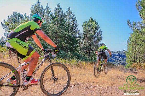 Dois ciclistas em bicicleta de gravel num caminho de terra entre pinheiros, equipados com coletes de neoprene amarelo e preto.