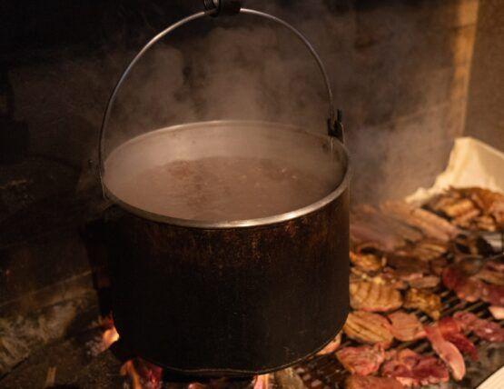 Caldeira de metal escuro suspensa sobre fogo, com vapor a sair, rodeada de lenha acesa.