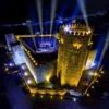Castelo medieval com torres de pedra iluminadas a amarelo e azul, com plateia e palco montados no pátio interior à noite.
