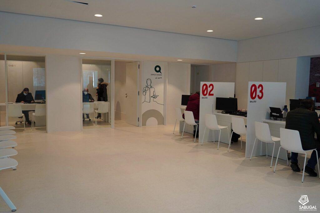 Interior de espaço de atendimento com balcões numerados 02 e 03, vidros divisórios e pessoas aguardando